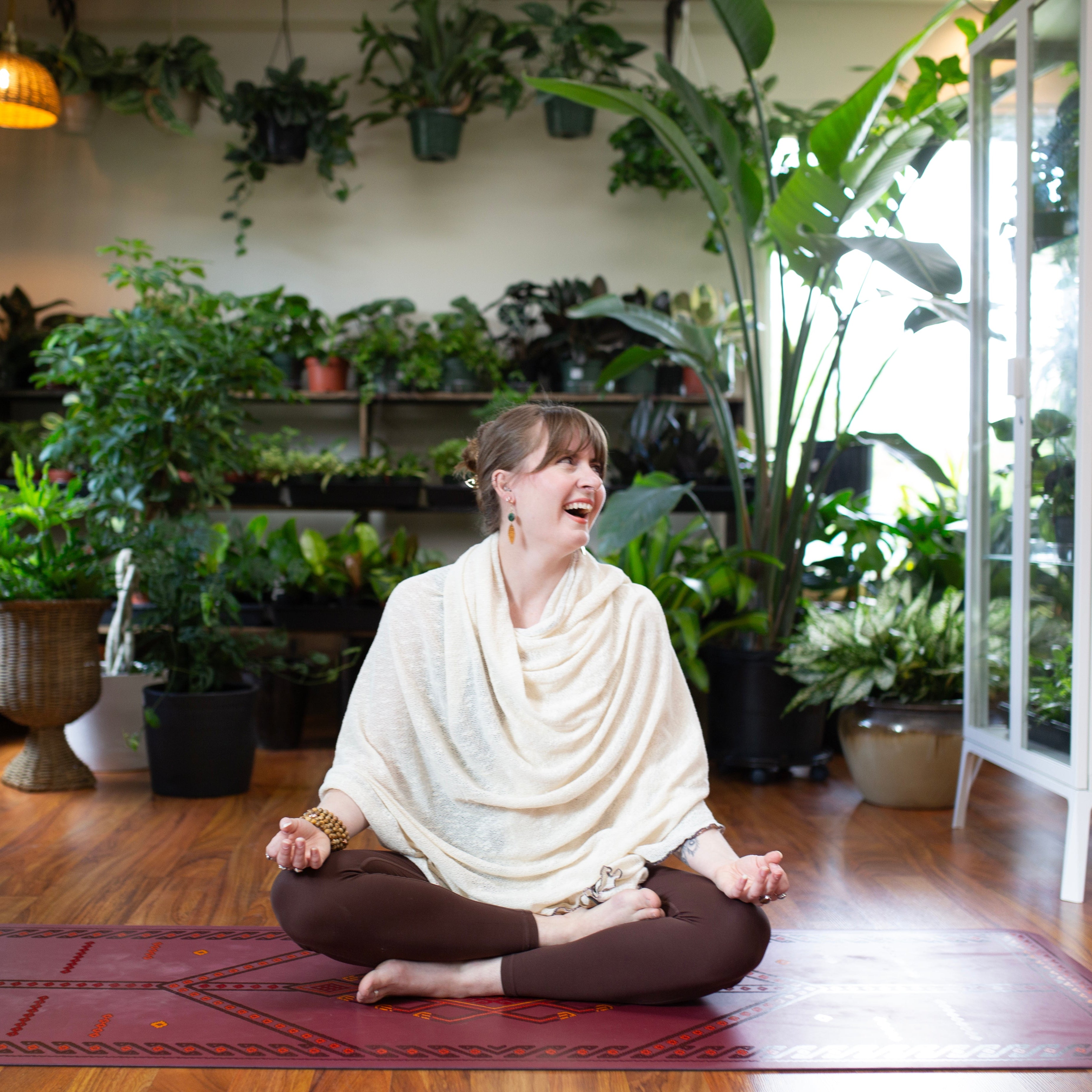 Cassidy in a white shawl and brown pants sitting in a meditative pose on a red mat surrounded by plants.