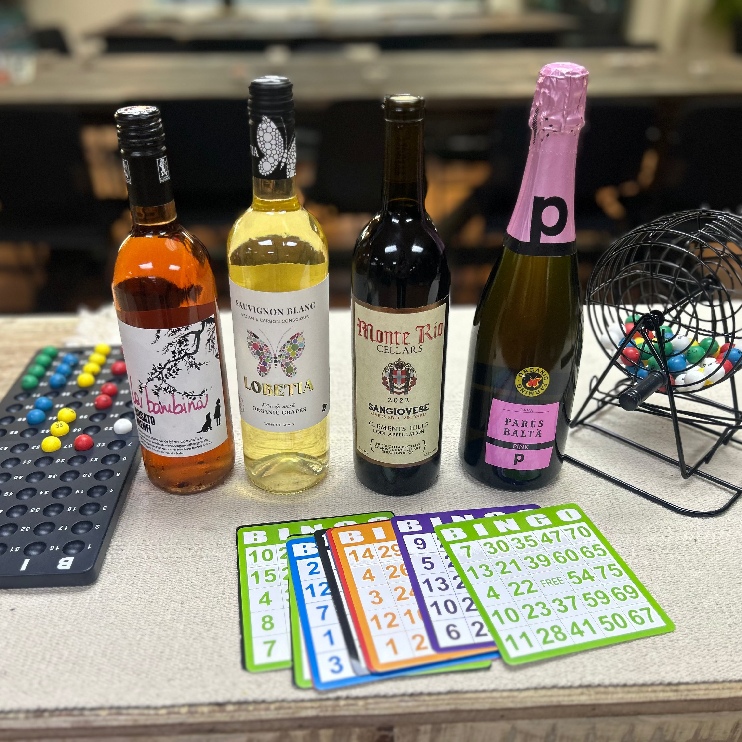Four bottles of wine on a table with bingo cards and a bingo roller in the background.
